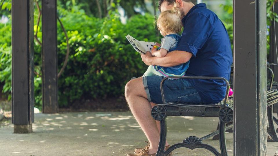 Bankta oturup kucağındaki çocuğa kitap okuyan baba